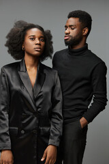A stylish African American couple poses elegantly in black attire against a neutral gray background, conveying confidence and contemporary fashion