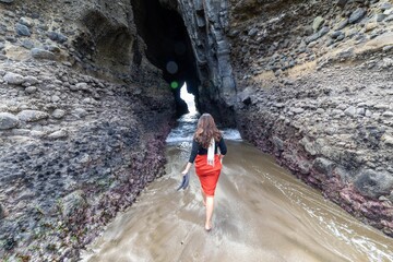 A woman walks barefoot through a sea cave in Piha, Auckland, New Zealand. She carries her shoes as...