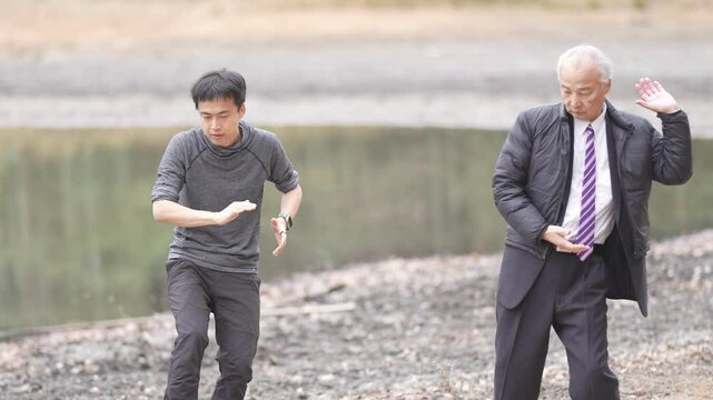 In a bamboo grove and pond park, a man in his 70s in a suit and a man in his 30s practice tai chi. Their slow movements are captured in a slow-motion video against the peaceful natural background.