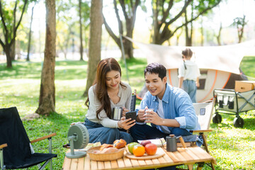 Asian couple enjoys a peaceful camping morning with coffee, breakfast, music, and devices in nature with love