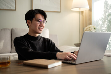Young man in glasses typing on a laptop while seated on a sofa in a cozy living room with a tea cup on the table.
