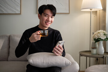 Cheerful man using a mobile phone while enjoying a hot drink in a living room.