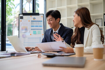 Professional man and woman discussing reports and smiling in a modern office space with charts, a laptop, coffee cups, and a large glass window in the background.