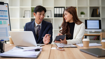 Businessman and businesswoman exchanging ideas during a meeting, surrounded by paperwork, technology, and presentation tools in a bright, contemporary office.