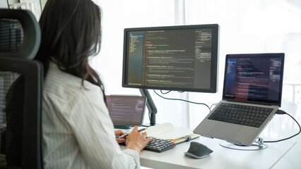 Rear view of a woman working on programming code with professional dual-monitor setup in bright...