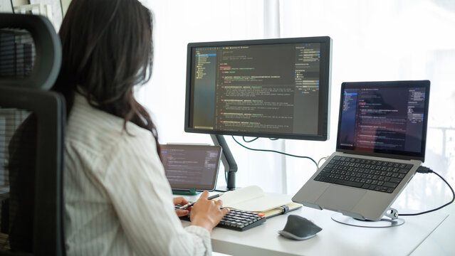 Rear view of a woman working on programming code with professional dual-monitor setup in bright workspace.