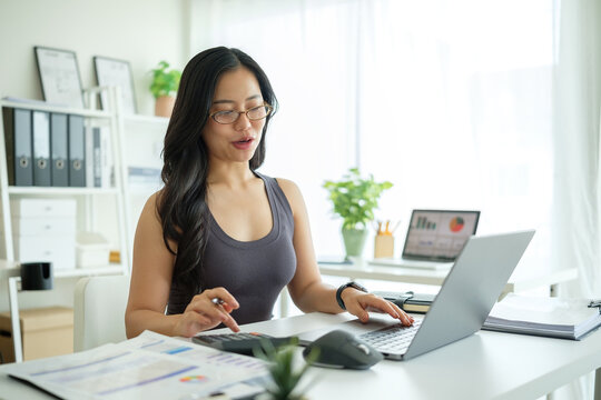 Asian woman reviewing financial data while working from home, surrounded by reports, charts, and digital devices.