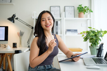 Smiling young woman holding stylus and digital tablet sitting in a bright home office.