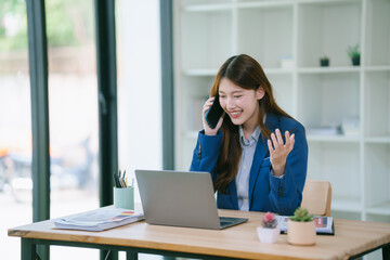 Asian businesswoman sitting talking on the phone at a desk full of documents, tablets, laptops planning her work.