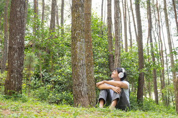 Happy young asian man wearing a white t-shirt and gray pants is sitting cross-legged on the grass in a lush pine forest while listening to calming music through his earphones.