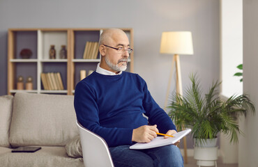 Serious senior man writing a book at home. Portrait of a bald bearded mature writer in spectacles sitting in a chair, holding a pencil and a notebook, looking away and thinking