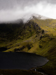 Misty mountain peak dramatically overlooks a dark, still lake, creating a serene yet powerful landscape. The contrast between the light and dark is breathtaking.