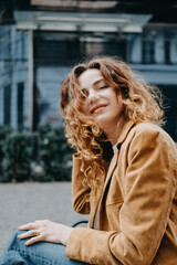 A young woman with curly red hair sits quietly on a bench looking down with a calm expression....