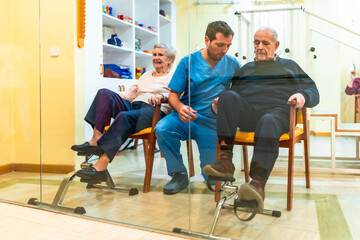 Nurse assisting elderly man and woman exercising with pedal exerciser in nursing home