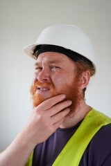 A construction worker with a white hard hat and a bright yellow safety vest contemplates, touching his red beard. His expression shows curiosity and thoughtfulness.