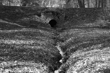 The black hole of an old brick drain with a small stream dividing the ravine in half in black and white style