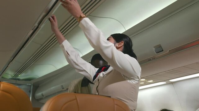 Friendly flight attendant. Air hostage helping passenger businessman put luggage on top shelf or cabin compartment on airplane, woman help black man lift suitcases putting overhead locker on airplane