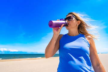 Hydration of body. Portrait of middle aged modern woman drinking water from water bottle on sandy beach on sunny summer day. Break from sports training to replenish water in body