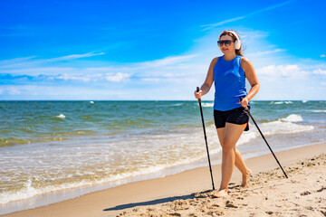 Playing sports on beach in summer. Pretty mature woman practicing Nordic walking and listening to...