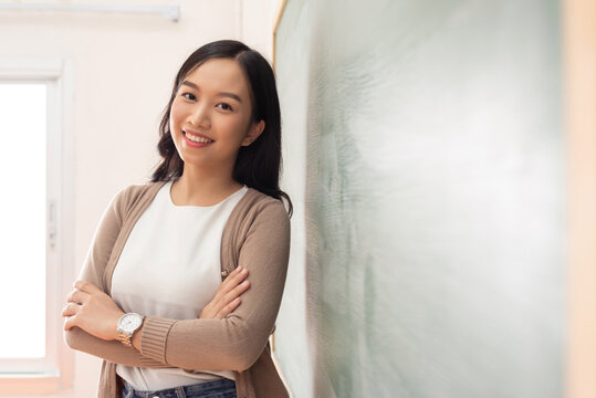 Smiling young Asian woman Student or teacher stands confidently with crossed arms next to a chalkboard in front of classroom
