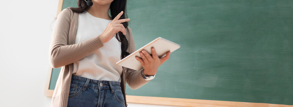 Asian woman stands confidently in front of green chalkboard of online classroom, holding digital tablet and gesturing explaining something with a thoughtful expression and hand gesture.