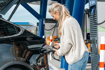 Blonde European woman smiling while connecting her electric car to charger green energy concept for...