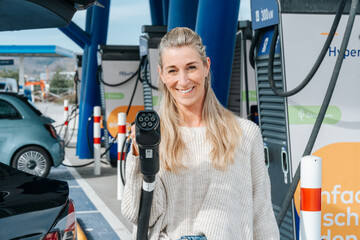 Happy blonde woman holds electric car charger in hand smiling at camera concept of green energy and...