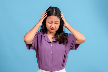 Young Asian woman with pained and frustrated expression, eyes closed, hand holding her head, standing against blue background. Showing discomfort, confusion, and stress naturally.