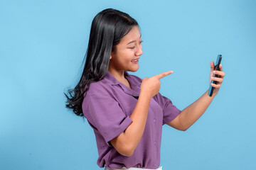 Cheerful young Asian woman pointing excitedly at a smartphone while smiling, showing a joyful and...