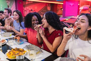 Young women sharing pizza slices with friends, enjoying a meal together at a bowling alley,...