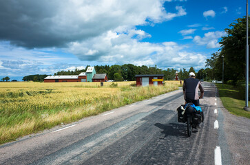 A cyclist with panniers rides along a rural road past golden wheat fields and a farm under a partly cloudy sky in the Swedish countryside, Västra Götaland County, Sweden