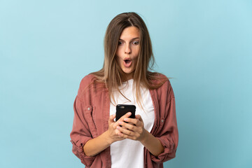 Fototapeta premium Young Slovak woman isolated on blue background looking at the camera while using the mobile with surprised expression