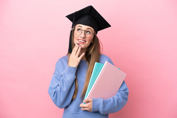 Young student woman wearing a graduate hat isolated on pink background looking up while smiling