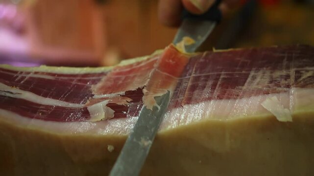 Close-up of a skilled hand slicing a prosciutto with a sharp knife, highlighting the precision and tradition involved in preparing this Italian delicacy.​