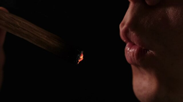 Close up of man exhales softly onto a glowing Palo Santo stick, releasing purifying smoke for spiritual cleansing, meditation, stress relief, and dispelling negative energy. Black background