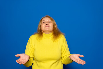 Maybe I have no idea. Portrait of a confused Caucasian woman in her 40s, shrugging her shoulders in bewilderment, hesitating and unsure, not knowing the answer. Isolated on a blue studio background.