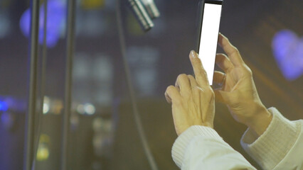 A close-up of the hands of a woman at a concert holding a smartphone and recording the performance, standing near the stage. White screen template