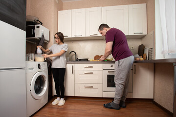 Man and woman cooking together in small kitchen, mixing dough and chopping vegetables.