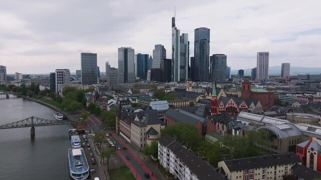 Aerial view of Frankfurt, Germany, featuring the Main River, Eiserner Steg, modern skyline, moving boats, vehicles, and lush greenery under an overcast sky.