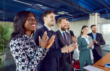 Group of diverse young people applauding standing at the desk in a row. Smiling successful coworkers and company employees clapping a colleague on a meeting or business conference.