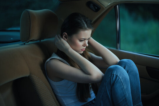 A troubled young woman sitting in a car's backseat, covering her ears, portraying emotions of distress and isolation, with a moody dark background.