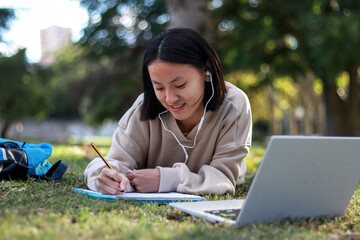 Young Asian woman lying on grass, taking notes while using a laptop