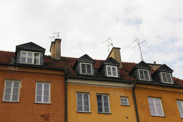 Fototapeta premium Row of traditional attic dormer windows and old-fashioned TV antennas on aged orange facades with red tiled roofs — classic European architecture under cloudy sky