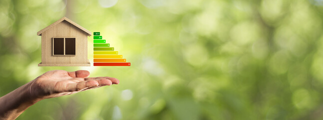 Hand holding wooden house icon with energy efficiency rating bars on blurred green background,...