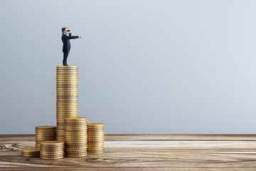 Businessman looking ahead with binoculars while standing on tall stack of coins, symbolizing...