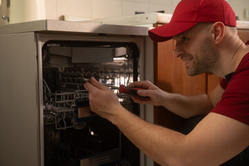 Professional maintenance worker wearing red uniform carefully diagnosing and repairing dishwasher inside residential kitchen, using specialized tools and expertise