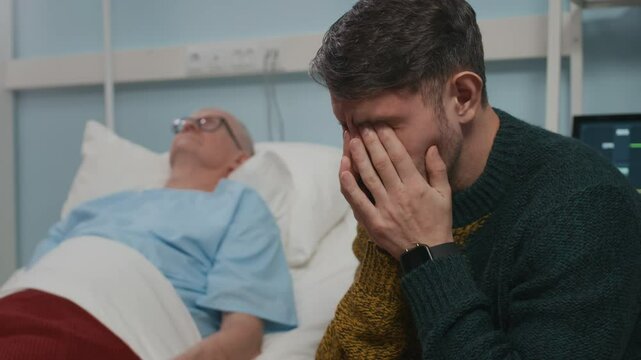 Focus on sad adult Caucasian son sitting by bed of sick senior father undergoing treatment in hospital room