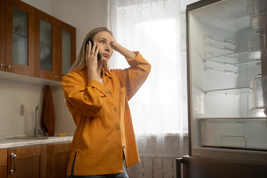 Frustrated woman in orange shirt talking on mobile phone, gesturing towards empty refrigerator in bright kitchen, reporting malfunction to customer service