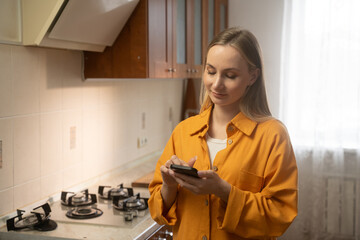 Woman wearing an orange shirt stands in her kitchen, using a smartphone app to select a gas stove repair technician, demonstrating the convenience of modern technology for home maintenance