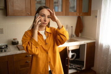 A woman in a yellow shirt stands in her kitchen while discussing urgent plumbing issues with a repair service regarding a leaky sink that needs immediate attention.
